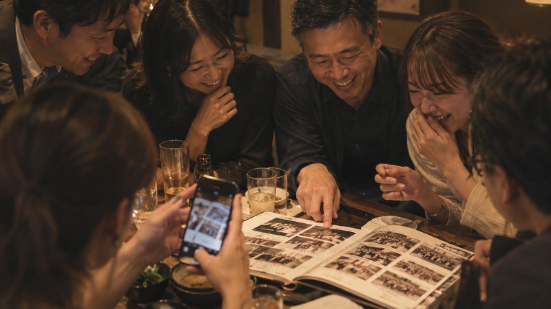 卒業アルバムを囲んで笑う同級生たち。1人がそのページをスマホで撮っている
